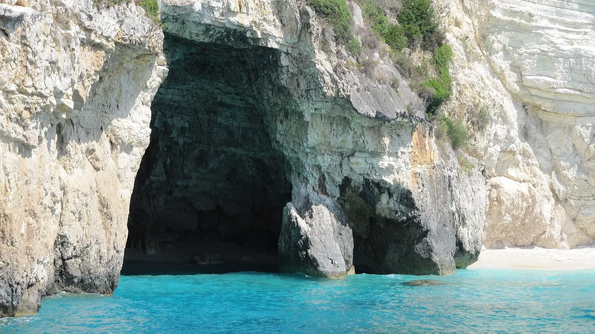 Sea cave mouth in limestone cliffs with turquoise water on a Greek island coast, the kind of cavern swimmers visit on Naxos boat tours to Rina Cave